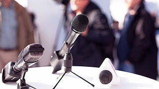 Close-up of two microphones, with the blurred image of a man in suit and tie in the background